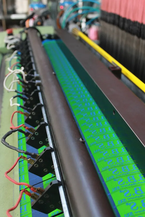 Woven label tape with lettering on a green ground being woven on the loom